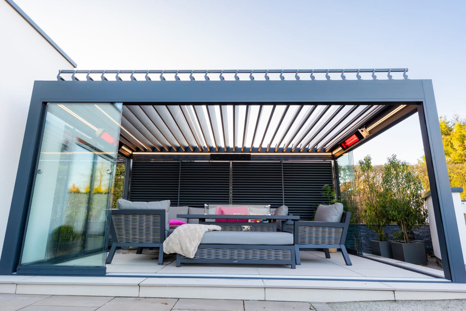 Aluminium pergola with a glass sliding door open onto a lounge and dining area enclosed by a louvred side screen