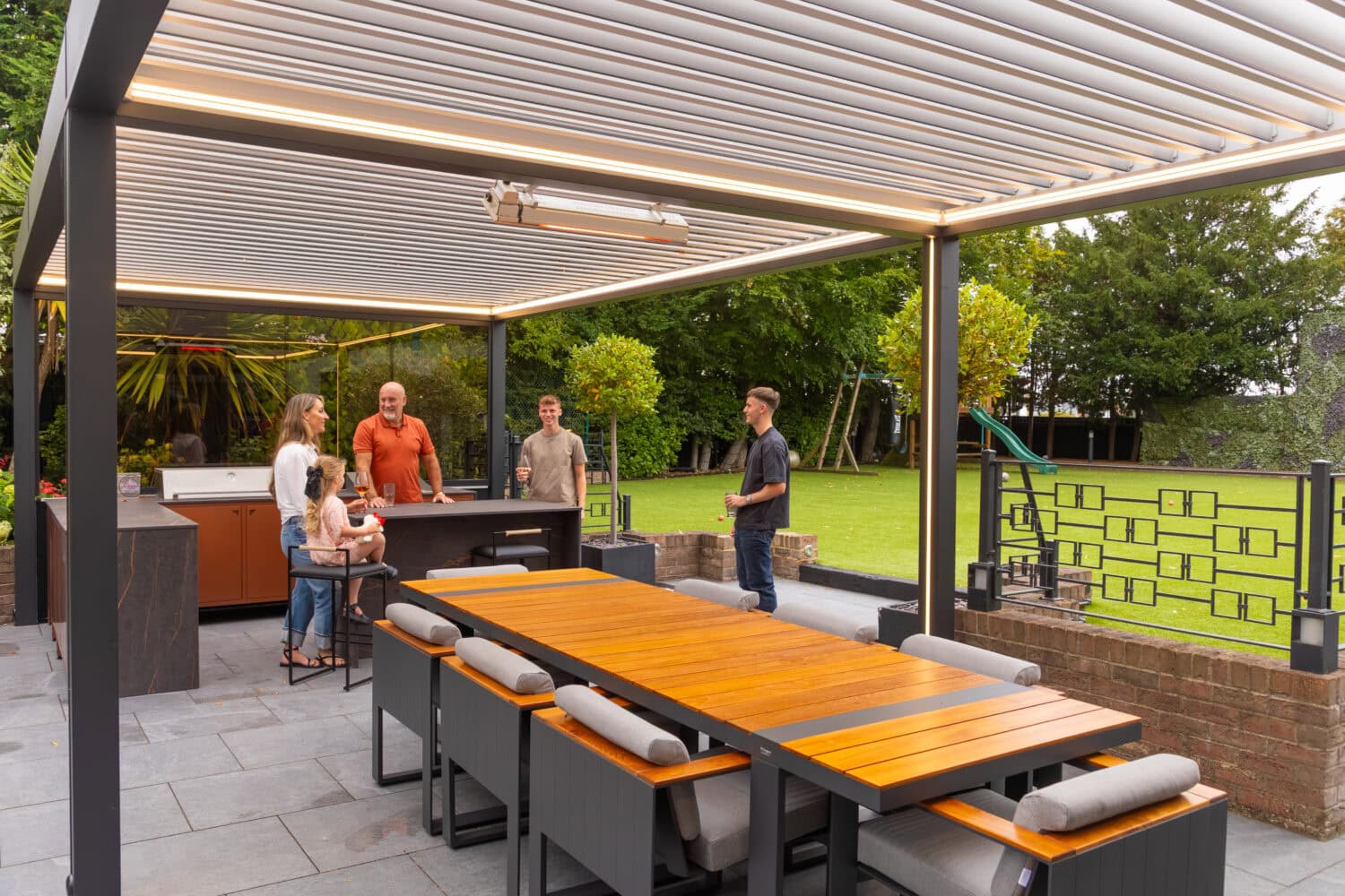 Family and friends gathered for an evening meal beneath an aluminium louvre roof pergola with an outdoor kitchen and warm integrated lighting