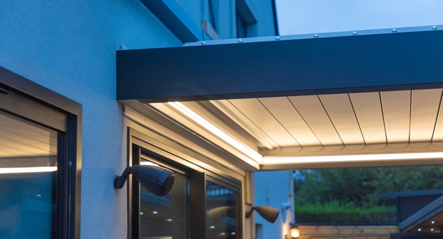 Detail of an aluminium pergola roof edge with integrated guttering and warm LED lighting at dusk