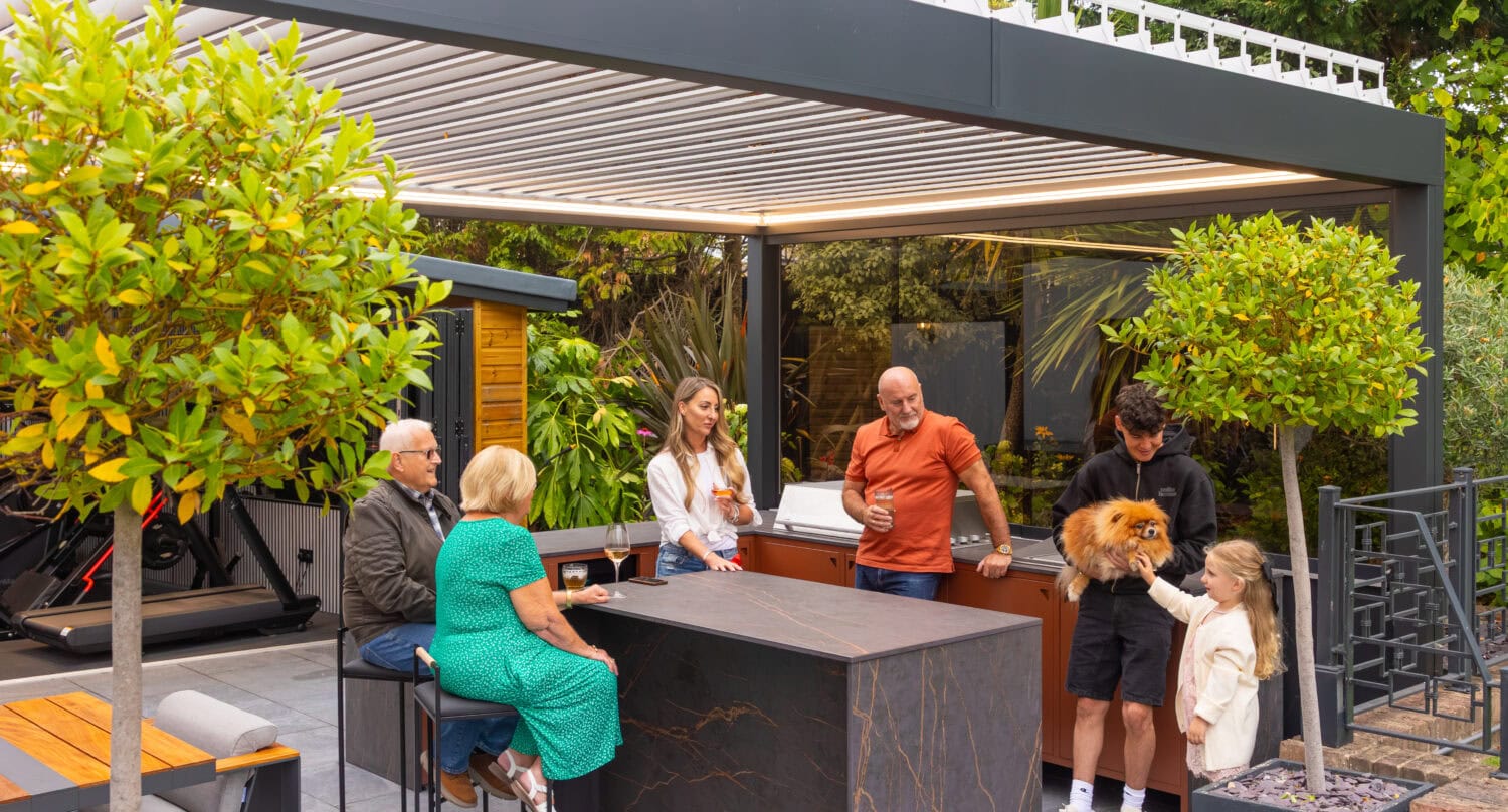 Family relaxing and dining beneath an aluminium louvre roof pergola with a built-in bar and outdoor kitchen
