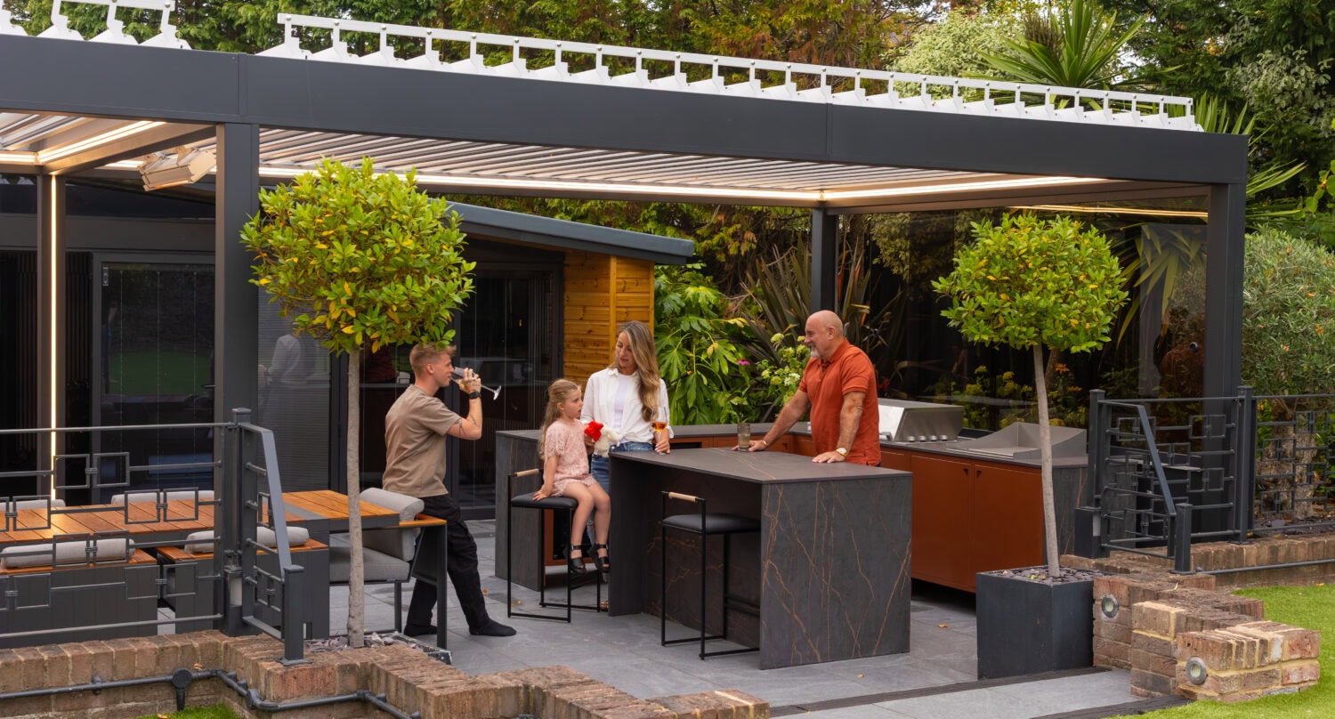Family with a dog gathered around an outdoor kitchen island beneath an aluminium pergola